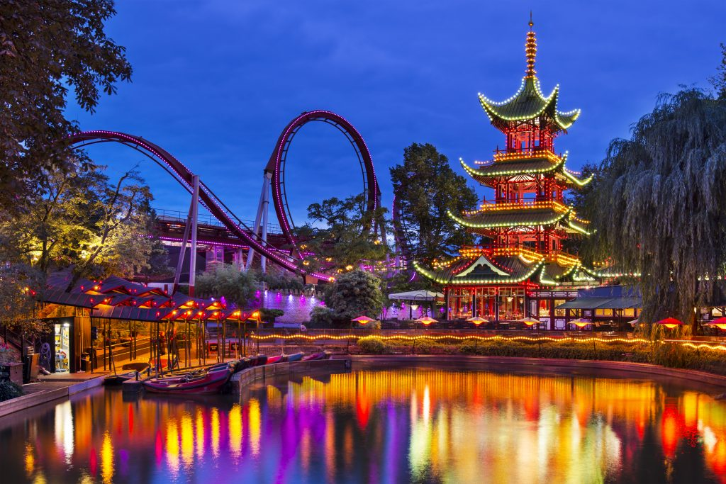 An amusement park at dusk with a lit pagoda, roller coaster loop, and glowing reflections on the water.