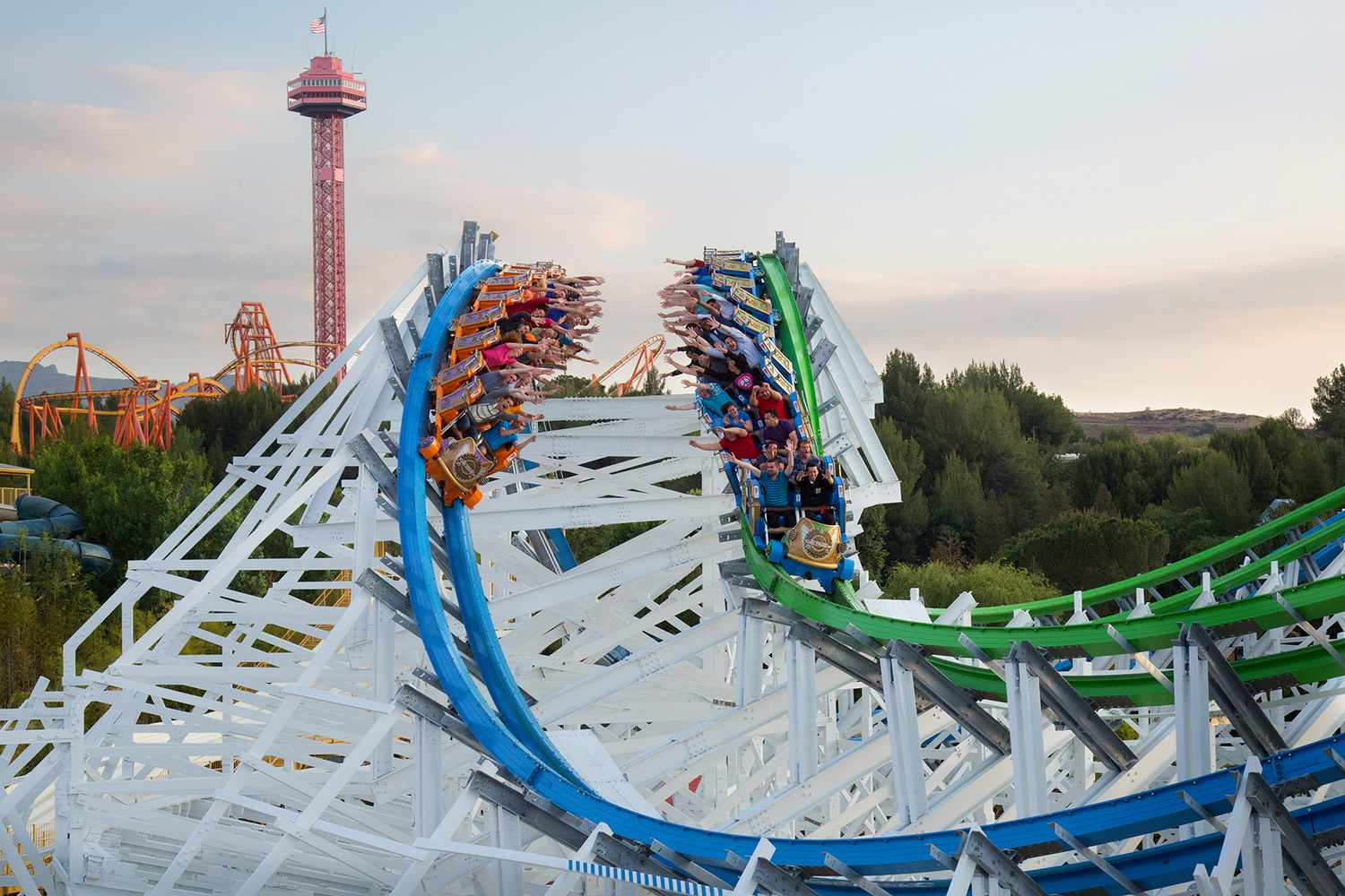 A blue-and-green dueling wooden coaster racing on white lattice track with a tower in the background.