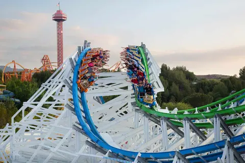 A blue-and-green dueling wooden coaster racing on white lattice track with a tower in the background.
